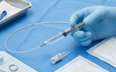 A clinician's blue-gloved hand holding a syringe drawing medication from a small glass ampoule on a blue sterile drape, surrounded by sterile packs and forceps.