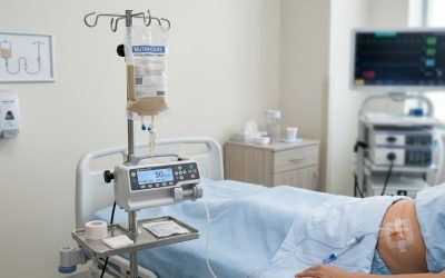 A patient lying in a hospital bed receiving enteral nutrition through a PEG feeding tube, with a Nutri-Care formula bag hanging on an IV stand connected to a feeding pump and tubing running to the patient's abdomen.